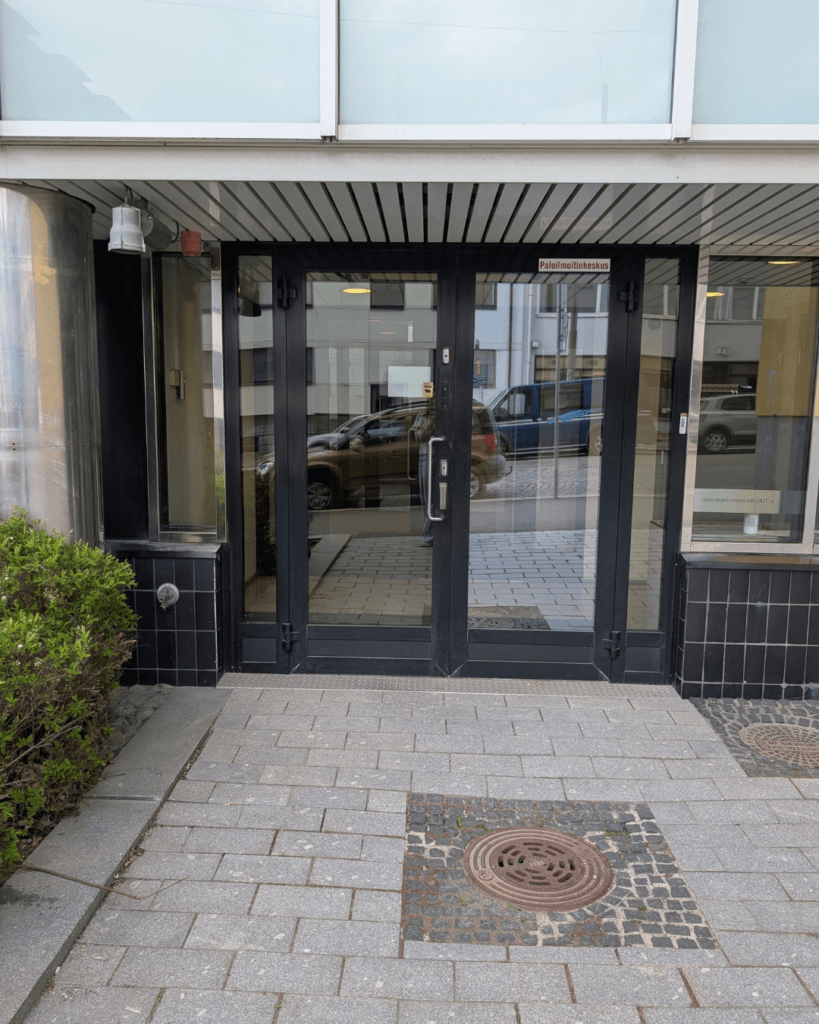 Rakennuksen sisäänkäynti katutasossa: tummat lasiset kaksoisovet, tasainen kivetty kulkuväylä ovelle ja ovessa vedettävä kahva.