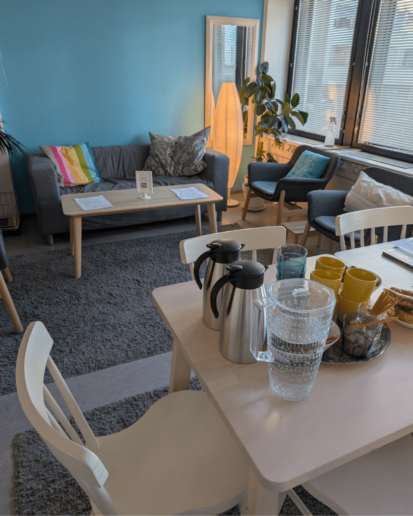 Kuvassa näkyy osittain UUT:n vertaistukihuone huoneen nurkasta ruokapöydän takaa kuvattuna: kaksi huonekasvia, kahden istuttava harmaa sohva, kaksi harmaata nojatuolia, värikkäitä tyynyjä, nurkassa paperivalaisin, jossa lämmin valo, lattialla harmaat karvamatot, sohvapöytä, jolla on papereita ja vaalean ruokapöydän kulma sekä muutama tuoli. Pöydälle on katettu kahvitarjoilut.